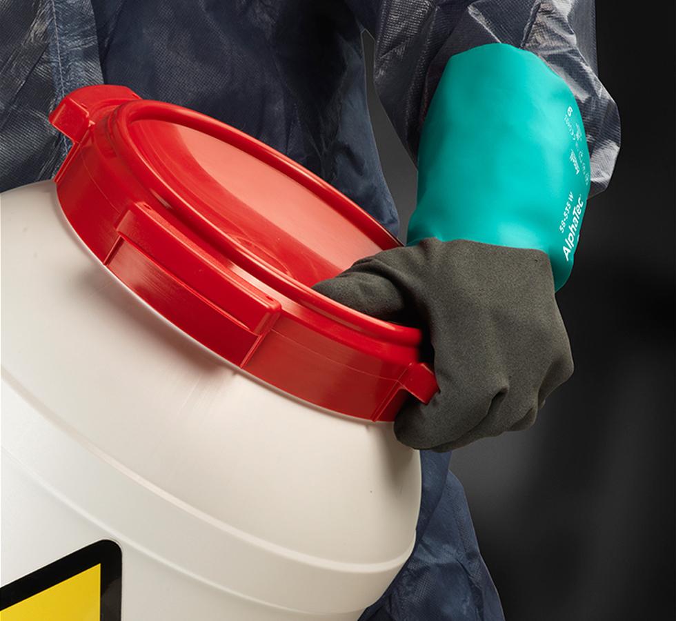 A Person In a Dark Coverall Wearing a Pair of Dark Grey and Blue Long Length Cuff ALPHATEC® 58-535B Nitrile Gloves Holding a Warning Container with a Red Lid - Sentinel Laboratories Ltd