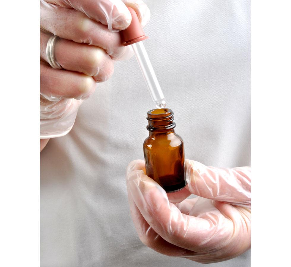 A Person Wearing a Wedding Ring and a White Lab Coat Using a Pair of Clear Shield GN65 Powder Free Vinyl Examination Gloves Squeezing Liquid from a Pipette into a Brown Vial  - Sentinel Laboratories Ltd