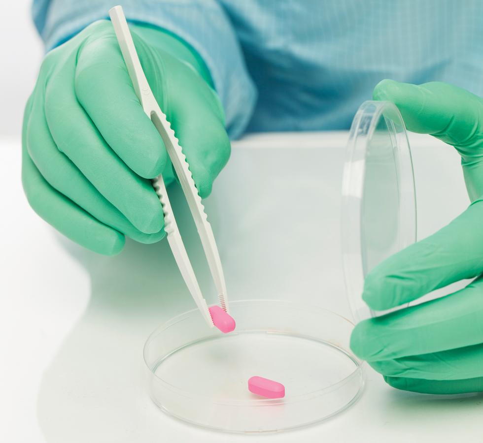 A Person in a Blue Lab Coat Wearing Green BioClean Fusion™ Non-Sterile 300mm Length Polychloroprene Gloves using Tweezers to Pick Up a Pink Pill - Sentinel Laboratories Ltd